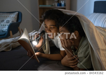 Positive African American girl shining flashlight on book while reading it to brother in darkness Positive African American girl shining flashlight on book while reading it to brother in darkness 84343162