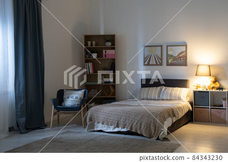 Empty bedroom with electric lamp on shelves, cozy armchair, pictures above bed at night Empty bedroom with electric lamp on shelves, cozy armchair, pictures above bed at night 84343230