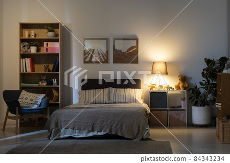 Cozy bedroom interior at night: stripped bed linen, nature pictures on wall, wooden shelves and switched lamp on bedside table Cozy bedroom interior at night: stripped bed linen, nature pictures on wall, wooden shelves and switched lamp on bedside table 84343234