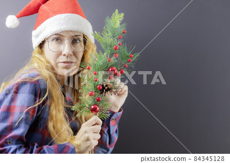 A blonde lady with glasses and a Santa hat holds an elegant spruce branch. Christmas poster A blonde lady with glasses and a Santa hat holds an elegant spruce branch. Christmas poster 84345128