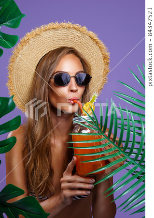 Attractive girl drinks fruit cocktail in sunglasses and straw hat while stands near tropical plants  84347521