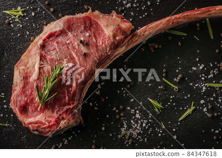 Raw beef tomahawk steak fillet with ingredients like sea salt, pepper, onion and herbs on black board, product image for restaurant 84347618