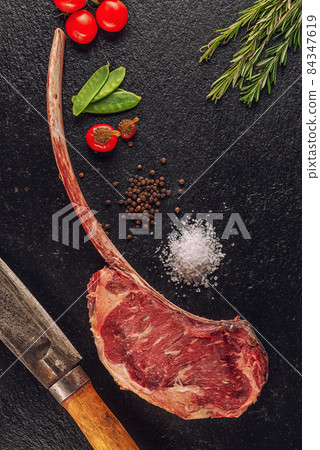 Raw beef tomahawk steak fillet with ingredients like sea salt, pepper, onion and herbs on black board, product image for restaurant 84347619