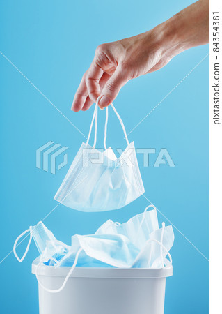 Hand throws a used face shield into the trash can on a light blue background. 84354381
