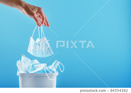 A hand throws a used protective mask into a trash can filled with masks from a virus on a blue background. A hand throws a used protective mask into a trash can filled with masks from a virus on a blue background. 84354382
