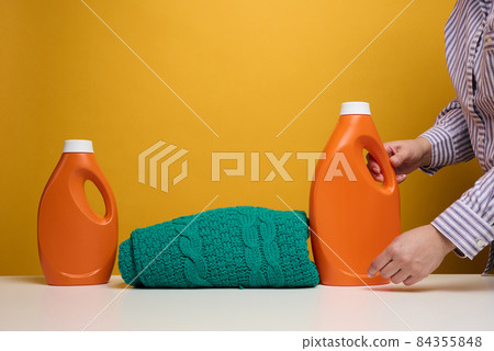 washed folded clothes and plastic orange large bottles with liquid detergent stand on a white table, yellow background. Routine homework 84355848
