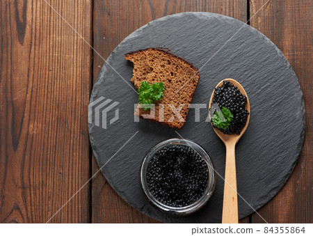 black paddlefish caviar in a wooden spoon, a slice of bread and a jar on a black round graphite board, top view 84355864