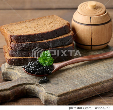 fresh grainy black paddlefish caviar in brown wooden spoon on a brown table, close up fresh grainy black paddlefish caviar in brown wooden spoon on a brown table, close up 84356068