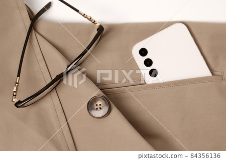 Modern smart phone in cream colour and glasses in the pocket of beige trench coat 84356136