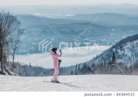 Woman taking photo on ski vacation using phone - Woman skier using phone app on ski trail slope taking pictures of amazing winter nature landscape. Girl using mobile smartphone in awesome ski wear Woman taking photo on ski vacation using phone - Woman skier using phone app on ski trail slope taking pictures of amazing winter nature landscape. Girl using mobile smartphone in awesome ski wear 84364535