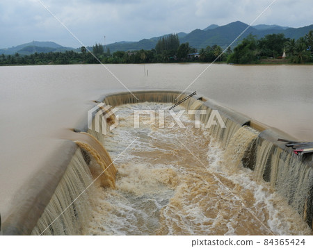 Brown water in the dam overflowing into the spillway with mountain and green forest in background , Flood in rainy season, Thailand 84365424