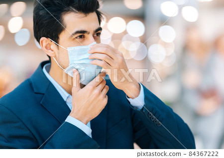Close-up side view of confident young man in stylish suit wearing protection face mask looking away in hall of mall centre, blurred background. Close-up side view of confident young man in stylish suit wearing protection face mask looking away in hall of mall centre, blurred background. 84367222