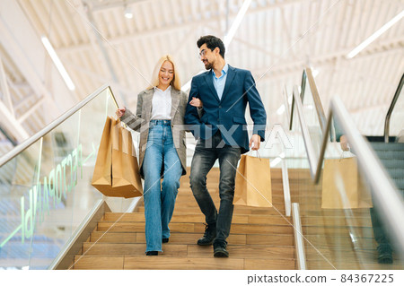 Low-angle view of smiling beautiful young couple holding shopping paper bags with purchases and walking down stairs, holding hands at mall. Low-angle view of smiling beautiful young couple holding shopping paper bags with purchases and walking down stairs, holding hands at mall. 84367225