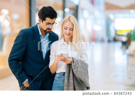 Portrait of happy beautiful young couple searching store in shopping mall using mobile phone. Front view of stylish man and woman buying at store. 84367243