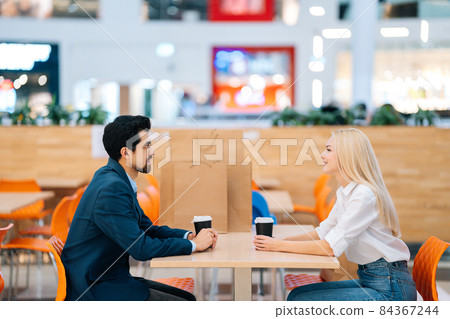 Handsome bearded man and attractive blonde woman having date sitting at table looking at each other in mall. 84367244