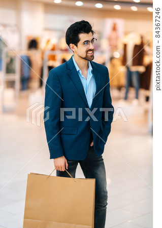 Vertical portrait of smiling elegance bearded young man in stylish suit and glasses holding shopping paper bags with purchases and looking away. Vertical portrait of smiling elegance bearded young man in stylish suit and glasses holding shopping paper bags with purchases and looking away. 84367246
