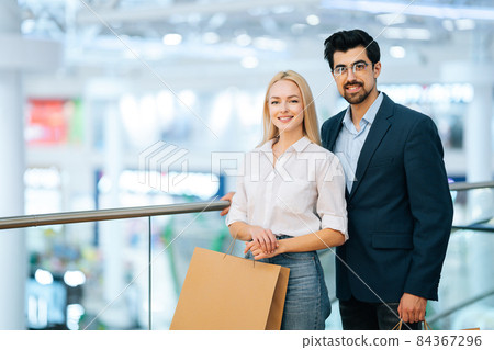 Beautiful young couple holding shopping paper bags with purchases and looking at camera, standing at mall. 84367296