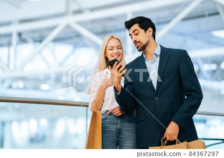 Cheerful beautiful young couple using phone holding shopping paper bags with purchases in mall, blurred background. H Cheerful beautiful young couple using phone holding shopping paper bags with purchases in mall, blurred background. H 84367297