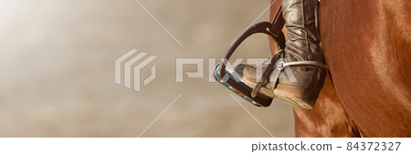 Foot of the athlete in a stirrup astride a horse, the foot of the rider, sitting on a horse, in a black boot with a spur, rests on a metal stirrup 84372327