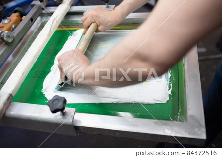 Serigraphy silk screen print process at clothes factory. Frame, squeegee and plastisol color paints 84372516