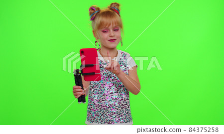Cheerful funny little teen girl kid dancing at camera filming video using phone on chroma key wall 84375258