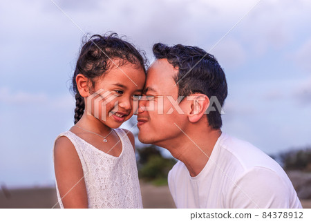 holidays, children and family concept -portrait of a little girl kissing her dad on cheek. Pretty girl giving a kiss to her father outdoor. Loving child embrace and kissing her father. 84378912