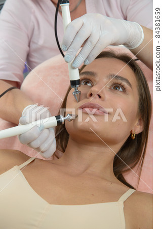 Beautiful woman at spa clinic receiving stimulating electric facial treatment from therapist. Closeup of female face during microcurrent therapy. 84381659