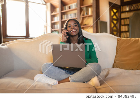 Smiling young black woman having conversation on mobile phone, working on laptop in living room, copy space 84382495