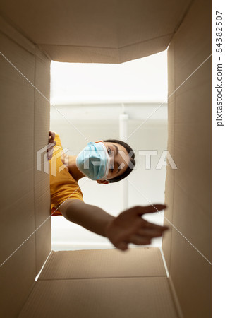 Young asian woman in medical mask unboxing cardboard parcel, trying to get something, view from the inside 84382507