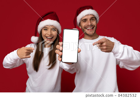 Xmas Ad. Happy Young Couple In Santa Hats Pointing At Blank Smartphone 84382532