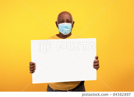 Senior black man wearing anti covid face mask, showing empty advertisement board over orange studio background, mockup 84382897