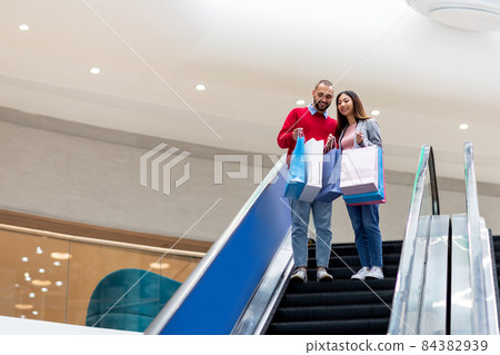 Full length of young interracial couple going down on moving stairway at mall, looking into shopping bags, empty space Full length of young interracial couple going down on moving stairway at mall, looking into shopping bags, empty space 84382939