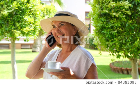 cute blonde senora woman, 50-55 years old, in a hat and a white dress, talks using a smartphone and cute blonde senora woman, 50-55 years old, in a hat and a white dress, talks using a smartphone and 84383093