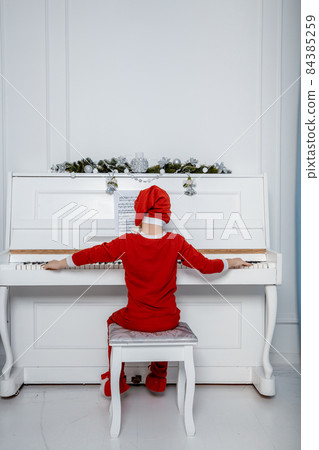 Boy in santa costume. Kid concept for merry Christmas and happy new year celebration. Small boy enjoys playing piano for the first time. Child play piano near the christmas tree. Christmas time. 84385259