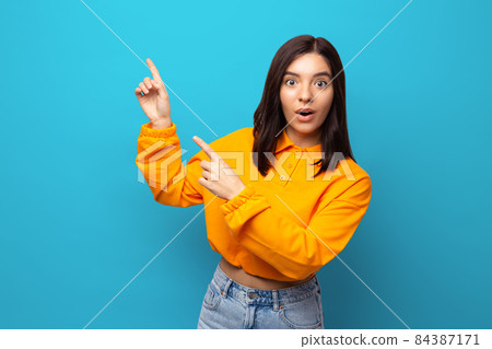 Beautiful multiethnicity woman in orange trendy shirt happily surprised expression pointing with fingers up against blue background Beautiful multiethnicity woman in orange trendy shirt happily surprised expression pointing with fingers up against blue background 84387171