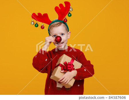 Cheerful little boy with wrapped Christmas gift holding red christmas tree ball in front of his nose 84387606