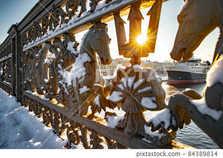 The view of the winter city Saint-Petersburg and big ship moored near English embankment through the railing of the bridge Blagoveshchensky, the lights of a sun, mooring lines, thick ropes The view of the winter city Saint-Petersburg and big ship moored near English embankment through the railing of the bridge Blagoveshchensky, the lights of a sun, mooring lines, thick ropes 84389814