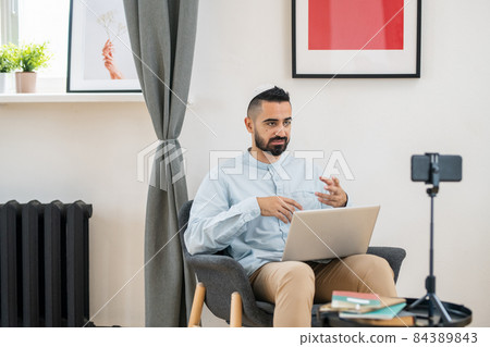Young bearded teacher or coach sitting in front of smartphone camera Young bearded teacher or coach sitting in front of smartphone camera 84389843
