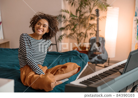 Cheerful African woman sitting near synthesizer at home Cheerful African woman sitting near synthesizer at home 84390103