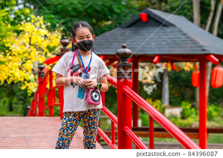 Asian child girl standing on wooden red bridge of Japanese traditional style, wearing face mask, holding a cup of fish food for feeding, hanging camera on her neck, daylight image Asian child girl standing on wooden red bridge of Japanese traditional style, wearing face mask, holding a cup of fish food for feeding, hanging camera on her neck, daylight image 84396087