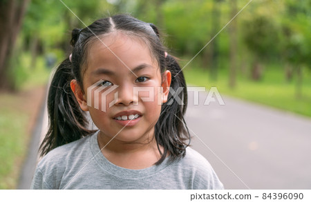 Portrait cute Asian child girl in the park, close up on the smiling face, some sweat drops on the face, gray color t-shirt, blurred background of trees in the park, sunlight image. 84396090