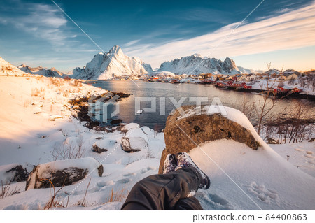 Legs of traveler relaxing in snow covered reine town on coastline at Lofoten islands 84400863
