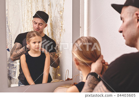 Cute little daughter and her tattoed dad are playing together near a mirror. Dad is doing his daughter's hair. Family holiday and togetherness. Cute little daughter and her tattoed dad are playing together near a mirror. Dad is doing his daughter's hair. Family holiday and togetherness. 84403605