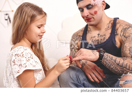 Funny time Tattoed father in a cap and his child are playing at home. Cute girl is doing makeup to her dad in her bedroom. Family holiday and togetherness Funny time Tattoed father in a cap and his child are playing at home. Cute girl is doing makeup to her dad in her bedroom. Family holiday and togetherness 84403657