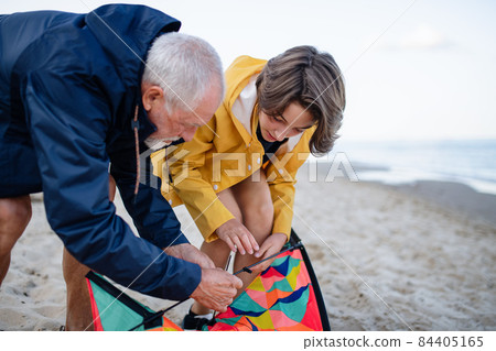 Senior man and his preteen granddaughter preparing kite for flying on sandy beach. Senior man and his preteen granddaughter preparing kite for flying on sandy beach. 84405165