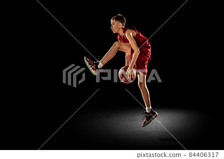 Full-length portrait of teen boy, basketball player in motion, training, dribbling ball under knee isolated over black background 84406317