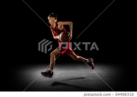 Full-length portrait of teen boy, basketball player in motion, training, running with ball isolated over black background 84406318