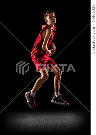 Full-length portrait of boy, teenager, professional basketball player scoring goal into jump, throwing ball into basket isolated over black background Full-length portrait of boy, teenager, professional basketball player scoring goal into jump, throwing ball into basket isolated over black background 84406320