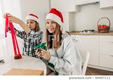 Two girls decorate cookies with bright red and green ribbons, prepare sweet treats for guests on Christmas or New Year's Eve Two girls decorate cookies with bright red and green ribbons, prepare sweet treats for guests on Christmas or New Year's Eve 84407212