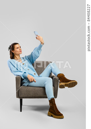 Middle age woman in blue warm knitted suit sits in armchair and making selfie Middle age woman in blue warm knitted suit sits in armchair and making selfie 84408227
